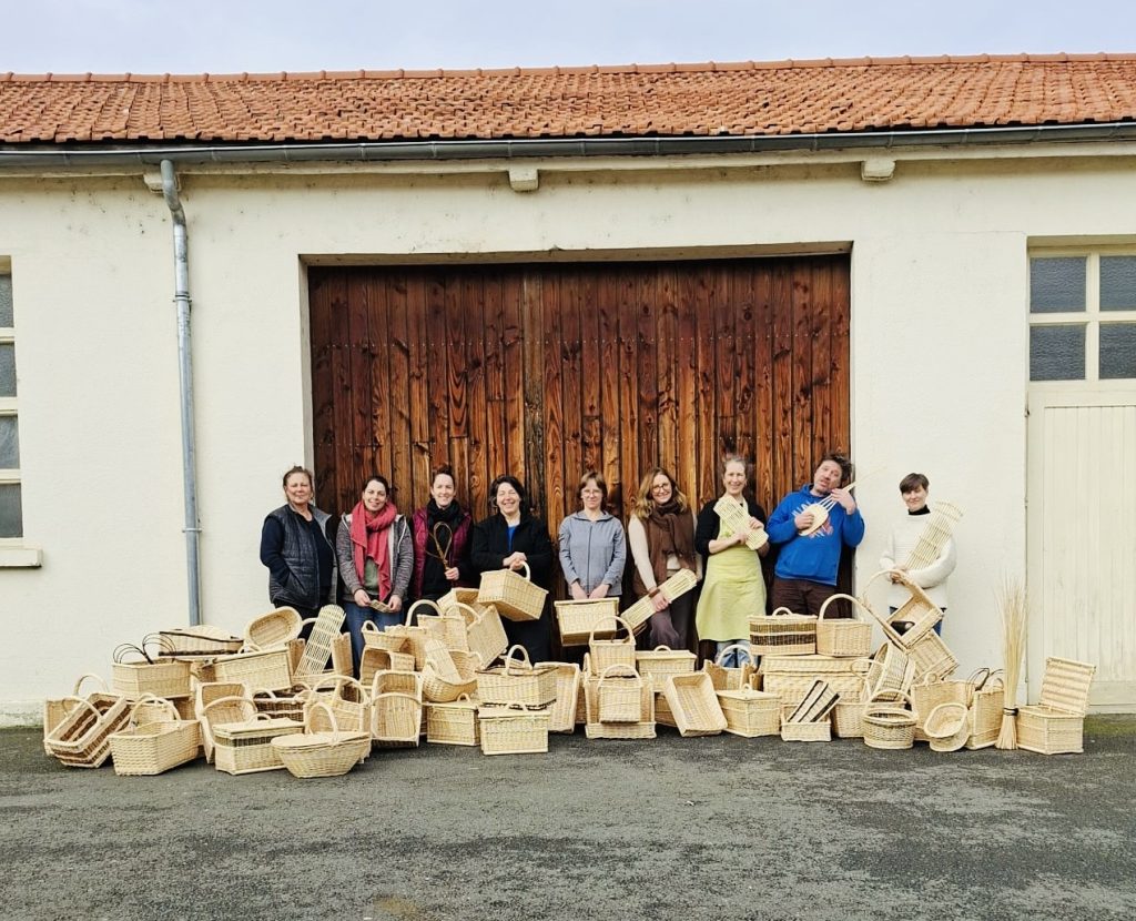 Groupe d'apprentis vanniers devant atelier avec l'ensemble de leurs productions en vannerie à leurs pieds.